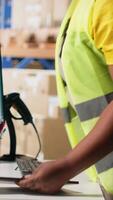 Vertical African american worker processing e-commerce cargo orders in warehouse setting, wearing hi vis vest and ensuring seamless parcel distribution. Cardboard boxes stored on big racks. Camera B. video