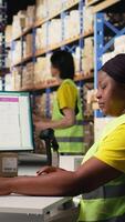 Vertical African american female worker typing awb shipment details on computer, updating package tracking software for clients. Person managing order fulfillment in a retail warehouse facility. Camera A. video