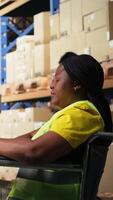 Vertical African american wheelchair user in safety vest processing cargo orders on pc at desk, logistics center optimized for reduced mobility. Female clerk handles online shopping operations. Camera A. video