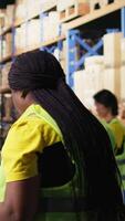 Vertical African american worker processing e-commerce cargo orders in warehouse setting, wearing hi vis vest and ensuring seamless parcel distribution. Cardboard boxes stored on big racks. Camera A. video