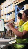 Vertical African american woman printing and applying airway bill tags on boxes, wearing hi vis vest in a warehouse near racks. Managing barcode labels with tracking info for order fulfillment. Camera A. video