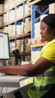 Vertical African american employee updating the order logistics software, preparing the shipment labels for distribution. Industrial depot staff with hi vis vest in e commerce logistics facility. Camera A. video