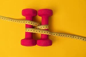 Concept of sports, fitness or weight loss. Two pink dumbbells for sports connected by a yellow tape measure on a yellow background with copy space. photo