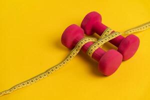 Fitness and diet concept. Pink dumbbells for sports connected by yellow tape measure on yellow background, side view with space for text. photo