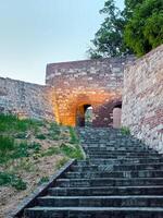 Stone steps lead to an illuminated archway in an old fortress. The path is flanked by historic brick and stone walls, leading the viewer into a serene twilight setting. photo