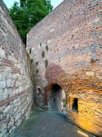Narrow medieval alley framed by ancient brick walls, featuring an arched tunnel and small window niche. Soft lighting enhances the aged texture of this fortress corridor photo