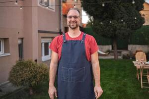 Smiling man in apron stands in a vibrant backyard during sunset gathering photo