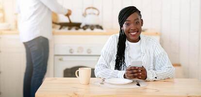 Young Black Woman Sitting At Table In Kitchen With Smartphone While Her Husband Cooking Lunch On Background, Millennial African American Couple Spending Time Together At Home, Selective Focus photo