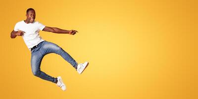 Positive black guy jumping up and showing copy space over orange studio background. Emotional african american millennial handsome man smiling and pointing at advertising or announcement photo