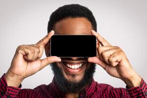 Look At This App. Black Guy With Smartphone Posing, Holding Phone Screen In Front Of Face Standing On White Studio Background. Man Covering Eyes With Cellphone Advertising Mobile Application. Mockup photo
