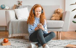Leisure Concept. Smiling Red-Haired Schoolgirl Sitting On The Floor Leaning On The Couch At Home, Listening To Music In Wireless Headphones And Chatting On Her Cell Phone, Enjoying Free Time photo