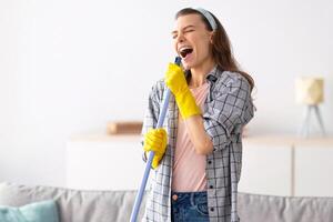 House cleaning is fun. Cheerful young lady using mop as mic, singing song while doing cleanup at living room. Millennial housewife pretending to be rock star while doing domestic duties photo