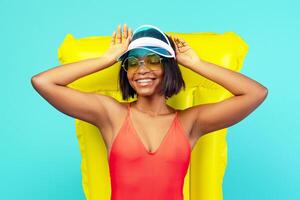 Summer vacation. Beautiful black woman in swimsuit lying on inflatable lilo, sunbathing on beach, blue studio background. Charming young lady relaxing and enjoying lazy day on pool float photo