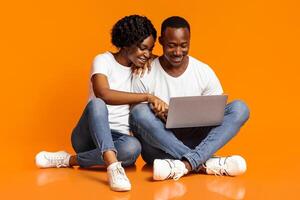 Relaxed african american man and woman using new laptop together while sitting on floor, checking social media, shopping online or watching photos, orange studio background, copy space photo
