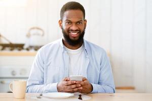 Young African Guy Sitting At Table In Kitchen With Smartphone While His Wife Cooking Lunch On Background, Black Millennial Couple Spending Time Together At Home, Selective Focus With Free Space photo