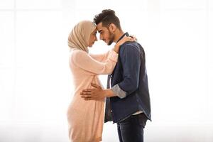 Romantic muslim spouses expecting baby standing together against window at home, loving arab husband tenderly touching belly of his pregnant wife in hijab, full length shot, side view with free space photo