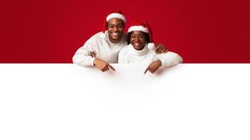Christmas list. Happy black spouses in santa hats leaning and pointing at white blank board with free space for you xmas advertisement or greetings, african couple standing on orange background photo