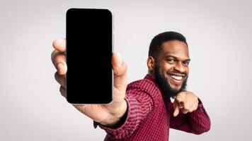 Use This Application. Cheerful Black Guy Showing Phone Empty Screen To Camera Recommending New Mobile App Posing Standing In Studio Over White Background. Smartphone Advertisement. Mockup photo