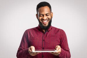 Cheerful African American Man Holding Digital Tablet Standing On White Background. Bearded Black Guy Playing Computer Game Online In Studio. Website, Internet Service And New Application Advert photo