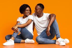 Happy african american lovers sitting on floor over orange studio background, looking at each other, copy space. Loving young black man and woman spending time together, bonding photo
