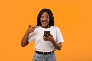 Excited black lady pointing at brand new smartphone in her hand, recommending new awesome mobile application, orange studio background, copy space. Smartphone, mobile phone, cellphone concept photo