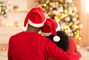 Young happy couple embracing and relaxing on comfortable couch. Loving young african american husband and wife in Santa Claus hats hug and look at Xmas tree and living room decor with garlands, back photo