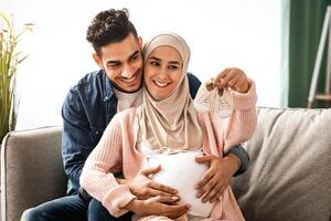 Portrait of happy pregnant muslim couple looking at tiny baby shoes while hugging on couch at home, young loving middle-eastern spouses embracing and enjoying future parenthood, free space photo