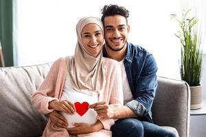 Happy Expectation. Cheerful Muslim Couple Holding Red Paper Heart Near Woman's Pregnant Belly While Posing Together On Couch At Home, Young Islamic Spouses Smiling At Camera, Free Space photo