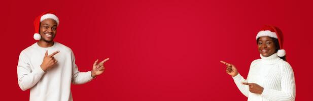 Excited african couple in santa hats pointing with fingers at free space in the middle of orange background, cheerful spouses showing empty place for your christmas offer or advertisement, panorama photo