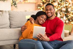 Modern new year, new normal and online during coronavirus quarantine. Millennial happy african american family use tablet and show credit card in living room with Xmas tree with glowing garlands photo