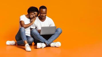 Carefree african american man and woman using new laptop together while sitting on floor, checking social media, shopping online or watching photos, orange studio background, copy space photo