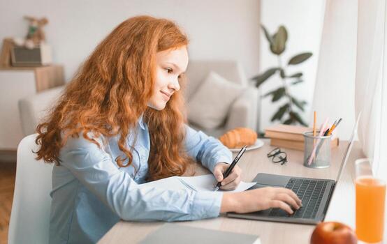 Lifestyle Concept. Portrait of positive red-haired generation z girl sitting at table, using laptop, typing on keyboard and while writing in her notebook, doing homework. Pupil having online class photo