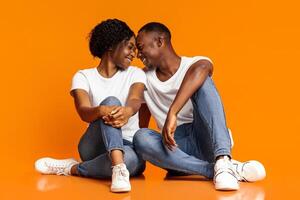 Emotional black lovers young man and woman cuddling on orange studio background, sitting on floor, touching each other with foreheads and laughing, enjoying time together, copy space photo