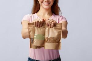 Eco-Friendly Package. Unrecognizable woman holding brown kraft paper doypack bags with weight food groceries in hands, lady buyer standing on grey background in studio, cropped image, closeup photo