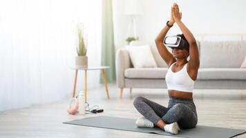 Black Woman Doing Yoga Wearing Virtual Reality Headset Training Sitting On Floor At Home. Smart Fitness, Digital Workout Via Augmented Reality Application Concept. Free Space For Text photo