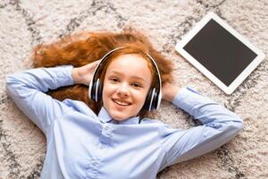 Rest And Relax. Top above high angle view of happy cheerful red-haired teenage girl listening to audio book, enjoying song wearing wireless headset, lying on floor carpet, looking at camera photo