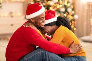 Free time together and new year at home during self-isolation. Laughing young african american guy and lady in Santa Claus hats hugging in living room with Xmas tree and decor with glowing garlands photo