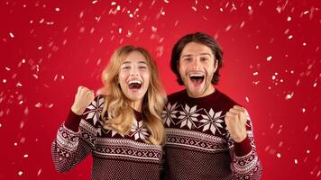 Excited young couple in Christmas sweaters screaming and making YES gesture on red background photo