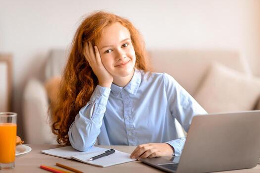 Distance Education. Portrait of smiling red-haired girl sitting at table, using laptop computer and looking at camera, doing homework, studying online at home. E-learning And Knowledge photo