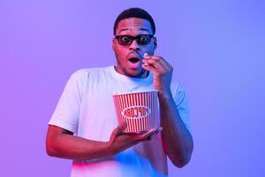 Portrait Of Shocked African American Guy In 3D Glasses Eating Popcorn From Bucket And Looking At Camera, Amazed Black Man Watching Movie In Cinema, Standing Under Neon light Over Purple Background photo