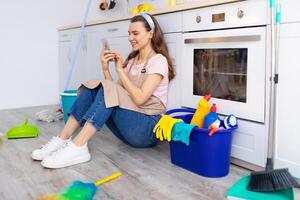 Happy young housewife taking break from home cleaning, browsing internet on smartphone while sitting on kitchen floor. Positive millennial lady checking social media during cleanup photo