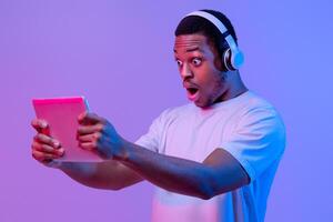 Cool Game. Excited African Man In Wireless Headphones Looking At Digital Tablet Screen, Amazed Black Guy Enjoying Modern Technologies For Gaming, Standing In Neon Light Over Purple Background photo