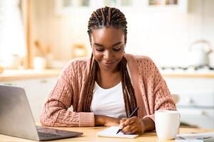 Smiling Black Student Woman Study With Laptop In Kitchen And Taking Notes, Young African American Lady Sitting At Desk At Home And Writing In Notepad, Enjoying Remote Distance Learning, Free Space photo