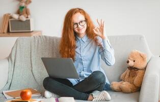Everything Is Okay. Positive smiling red-haired teenager girl in glasses showing ok sign gesture sitting on the sofa with crossed legs and using laptop, doing homework, recommending online school photo