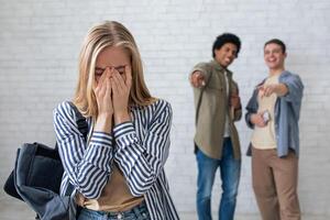 Bullying, negativity and peer problems. European and african american teens zoomers guys laugh at crying female with backpack in school or college, on white wall background, studio shot, empty space photo