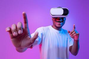 Augmented reality world. Excited black millennial guy in vr glasses, touching virtual button while playing game in goggles, standing in neon light over purple background, copy space photo