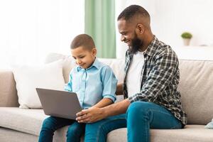 Happy Black Daddy Teaching Kid Son To Use Laptop Computer Sitting On Couch And Browsing Internet Together At Home. Technology And Gadget, Weekend With Father Concept photo