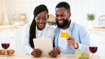 Cheerful Black Spouses Ordering Food Online With Digital Tablet And Credit Card, Sitting At Table In Kitchen, Purchasing Grocery Delivery, Enjoying Modern Technologies For Daily Life, Closeup photo