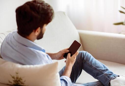 Unrecognizable Man Using Smartphone With Blank Black Screen While Relaxing On Couch At Home. Mockup Image With Bearded Man Messaging Or Shopping Online On Cellphone, Resting In Living Room, Copy Space photo