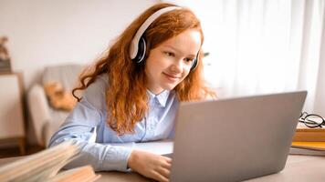 Online School. Closeup portrait of smiling red-haired girl using laptop, wearing wireless headphones, studying remotely, doing homework, watching movie or playing internet games photo